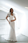 A woman modeling a white floor-length gown with dramatic feathered shoulder details and a long sheer veil, standing in a bright white architectural space.