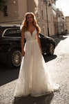 A woman wearing a sleeveless white lace wedding gown stands on a sunlit city street beside a black SUV, with buildings in the background.