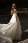A woman wearing a white wedding gown stands outdoors on a paved street, holding out the full train of the dress. The dress has a fitted bodice with lace detailing and a voluminous tulle skirt.