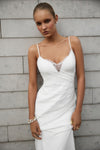 A woman modeling a white spaghetti-strap dress with a lace V-neckline, standing against a gray wood-paneled wall.
