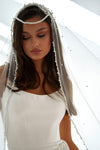 A woman wearing a bridal veil with pearl bead details and a matching headpiece, paired with a white wedding dress against a bright white background.