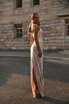 A woman stands outdoors on a paved street in front of a stone building, wearing a long sleeveless lace dress with a high slit and backless cutout. The dress appears light beige or ivory under warm sunlight.
