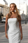 A woman wearing a strapless white textured tube dress stands on a city street, with blurred buildings and a vehicle in the background.