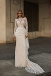 A woman modeling a long-sleeve ivory lace wedding gown with a fitted bodice, high neck, and a long train in an outdoor urban setting.
