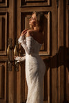 A woman wearing a fitted off-the-shoulder lace wedding dress stands in front of large dark wooden doors, with warm sunlight casting shadows on the wood and wall.