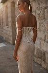 A woman wearing a strapless lace wedding gown with a lace-up corset-style back, photographed from behind in warm sunlight against a stone wall.