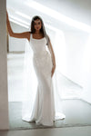 A woman wearing a sleeveless white wedding gown with a fitted bodice, draped overlay details, and a long sheer veil, standing in a bright modern interior with strong backlighting.