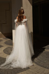 A woman wearing a voluminous white wedding gown with off-the-shoulder puff sleeves and a long sheer tulle train, photographed outdoors against a sunlit stone wall and patterned pavement.
