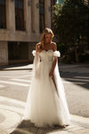 A woman wearing an off-the-shoulder white tulle wedding gown with a sheer draped overlay, standing on a city sidewalk in bright sunlight.