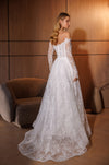 A woman wearing a white off-the-shoulder lace wedding gown with long sheer sleeves and a full skirt, standing in a warmly lit room with wood-paneled walls and a curved upholstered chair.