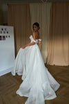 A woman wearing a white off-the-shoulder wedding gown with a full skirt, standing in a room with beige curtains and a wooden floor.