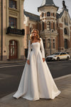 A woman wearing a long-sleeve white A-line wedding gown stands on a city street with ornate historic buildings and a car in the background.