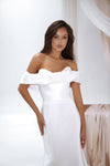 A woman modeling an off-the-shoulder white satin gown with a sweetheart neckline and a fitted silhouette, set against a soft white studio background with draped fabric.