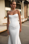 A woman modeling a strapless white wedding gown with a structured corset-style bodice, beaded waist detail, and a fitted mermaid-style skirt in an outdoor patio setting.