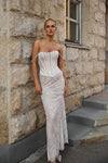 A woman stands outdoors against a stone building wall wearing a strapless fitted gown with a white corset-style bodice and a sheer pale pink lace skirt.