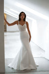 A woman modeling a white mermaid-style wedding gown with a lace-embroidered bodice and thin straps, standing in a bright, modern white interior space.