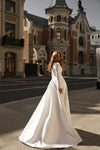 A woman wearing a long-sleeved white wedding gown with a high neck and structured back, standing on a city street in front of historic brick buildings.