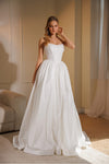 A woman modeling a strapless white ball gown with a structured corset-style bodice and a full flowing skirt, standing in a softly lit indoor room with beige walls and curtains.