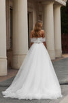 A woman wearing a white off-the-shoulder wedding gown with a lace bodice and full tulle skirt, photographed from behind in front of a building with large columns.