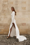 A woman wearing a long-sleeved white satin gown with a high thigh slit and a train, standing on a cobblestone street against a beige stone wall.
