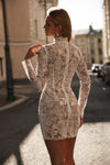A woman stands on a sunlit city street wearing a fitted long-sleeve lace mini dress with a high neck and back zipper. The dress is cream or off-white lace over a nude-toned underlayer, and the background shows blurred buildings and parked cars.