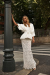 A woman posing outdoors in a white two-piece outfit consisting of a short-sleeve belted wrap-style top and a matching white lace maxi skirt, standing next to a dark street lamp post.