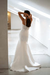 A woman wearing a strapless white mermaid-style wedding gown stands in a bright, modern interior with high windows and concrete walls, viewed from behind as she adjusts her hair.