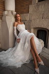 A woman wearing a strapless white lace wedding gown with long lace sleeves and a full tulle train, seated on stone steps beside a column in an outdoor architectural setting.