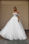 A woman wearing a voluminous off-the-shoulder white wedding gown with a fitted lace bodice and a full tulle skirt, standing in front of a beige paneled wall.