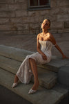A woman wearing a strapless white lace corset dress and matching pointed-toe heels sits on stone steps in front of a rustic stone wall.