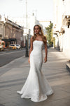 A woman wearing a strapless white lace mermaid-style wedding gown stands on a city sidewalk, with buildings and parked cars visible in the background.