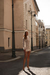 A woman stands on a sunlit city street wearing a fitted, long-sleeve mini dress with a textured, metallic champagne-gold pattern. The background features classic beige stone buildings and dark vintage street lamps.