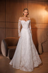 A woman models an off-the-shoulder white lace wedding gown with a fitted corset bodice and a full flared skirt, standing in a warmly lit room with wood-paneled walls and a curved upholstered chair.