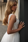 A woman in a sleeveless white lace and bead embellished wedding gown with a full tulle skirt, posing against a dark gray pillar.
