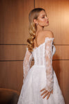 A woman wearing a white off-the-shoulder lace wedding gown with long sheer lace sleeves, photographed indoors against a warm wood-paneled background.