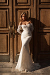 A woman wearing an off-the-shoulder lace wedding gown stands in front of large dark wooden doors, with warm sunlight casting shadows on the wall behind her.