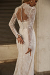A woman wearing a fitted lace wedding dress, shown from the back in a real interior setting with a stone wall and dark metal shutter visible.