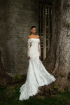 A woman wearing an off-the-shoulder lace wedding gown with long sheer sleeves and a fitted mermaid-style skirt stands outdoors beside a large tree trunk and stone wall.