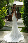 A woman wearing a white sleeveless wedding dress with lace and tulle detailing, photographed outdoors on a stone path with greenery and an umbrella in the background.