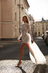 A woman walks down a sunlit city street wearing a fitted, long-sleeve mini dress covered in intricate floral lace, with a flowing sheer white train and nude heels.