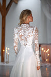 A woman shown from the back wearing a white bridal gown with a high neck, long sheer floral lace sleeves, and a full tulle skirt in a softly lit indoor setting.