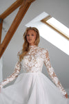 A woman modeling a white long-sleeve wedding dress with a sheer floral lace bodice and a full tulle skirt, posed indoors against a bright attic-style background with wooden beams and a skylight.