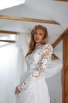 A woman wearing a white long-sleeve lace dress with floral embroidery and a sheer mesh bodice, standing in a bright white interior with wooden beams and draped fabric.