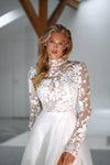 A woman modeling a white bridal gown with a sheer floral lace long-sleeve bodice and a flowing white skirt in a bright indoor setting.