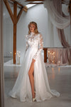 A woman modeling a long-sleeved white wedding gown with a high lace bodice, sheer sleeves, and a flowing tulle skirt with a thigh-high slit, photographed in a softly lit indoor venue with wooden beams and draped fabric.