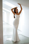 A woman modeling a sleeveless white mermaid-style gown with a fitted bodice, ruched waist, and slight train, standing in a bright white architectural interior with high windows.