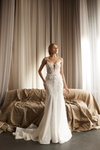 A woman wearing a fitted white wedding gown stands in front of draped beige curtains and a tan sofa. The dress has thin straps, a deep V neckline, lace applique, and a slight train.