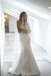 A woman wearing a white sleeveless wedding gown with a long sheer veil decorated with small floral appliqués, standing in a bright white hallway.