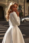 A woman wearing a white long-sleeve dress with structured shoulders and a flared skirt, photographed outdoors on a city street with blurred buildings and a car in the background.