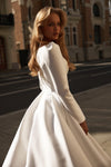 A woman wearing a white long-sleeve wedding dress with a structured bodice and full skirt, photographed outdoors on a city street with buildings in the background.